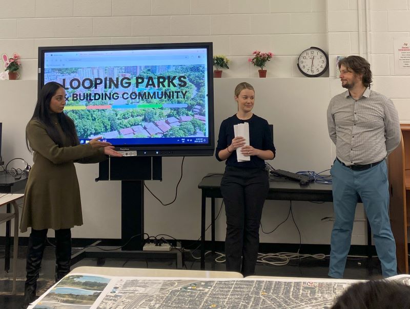 Three individuals, architects from the Western Loop project, stand in a classroom or meeting room with a large screen displaying the words "Looping Parks: Building Community" and an aerial image of a green park. A table with maps or architectural plans is visible in the foreground.