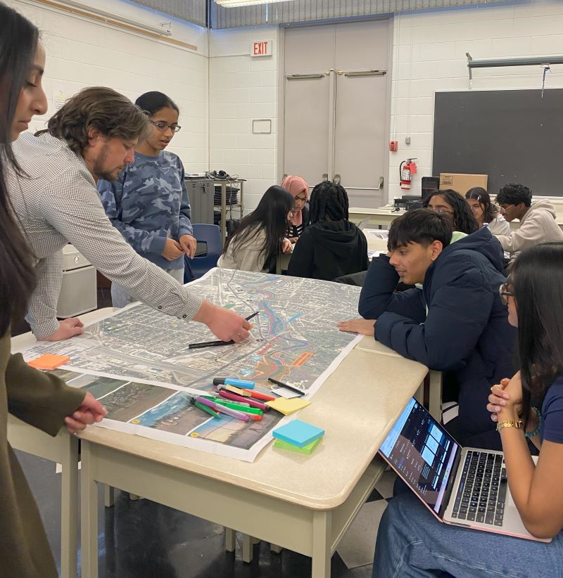 A group of students and two architects collaborate around a large table in a classroom. The table holds a detailed map with colorful markings, sticky notes, and highlighters. One of the architects leans over the table, pointing to a specific spot on the map while explaining.  Additional students in the background are engaged in similar group discussions.