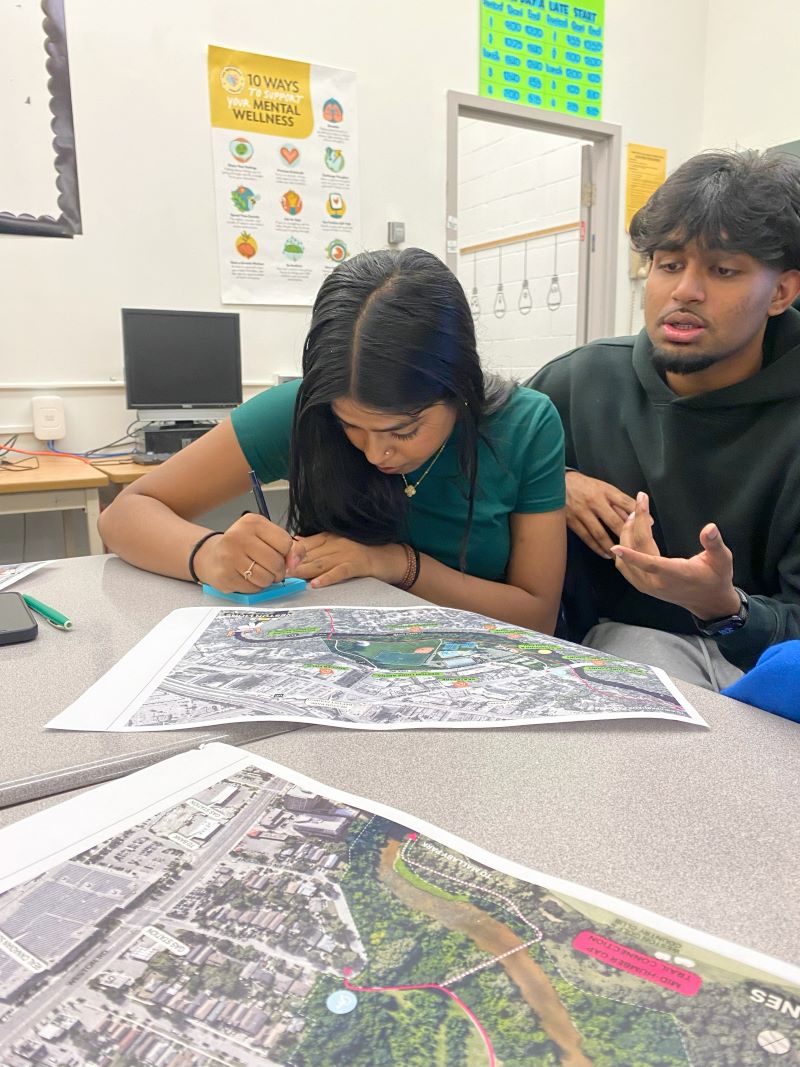 Two students sit at a table with a detailed map in front of them, one student is speaking and the other is taking notes on a piece of paper. 