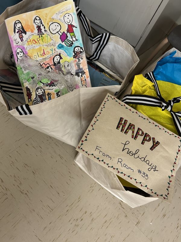 Two large gift bags filled with holiday items. One bag contains a colorful handmade card decorated with the text "Happy Holidays" in multicolored letters. The second bag displays a brown card reading "Happy holidays From: Room #33," adorned with festive light string designs around the edges.