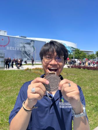 Male student is smiling standing in field holding bronze medal between two hands.