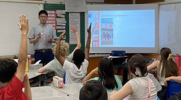 Image 1: City staff member stands at the front of a classroom with students sitting in desks with hands raised. On a projection screen there is an image from Chinatown Toronto with text “Ice Breakers” on the screen.