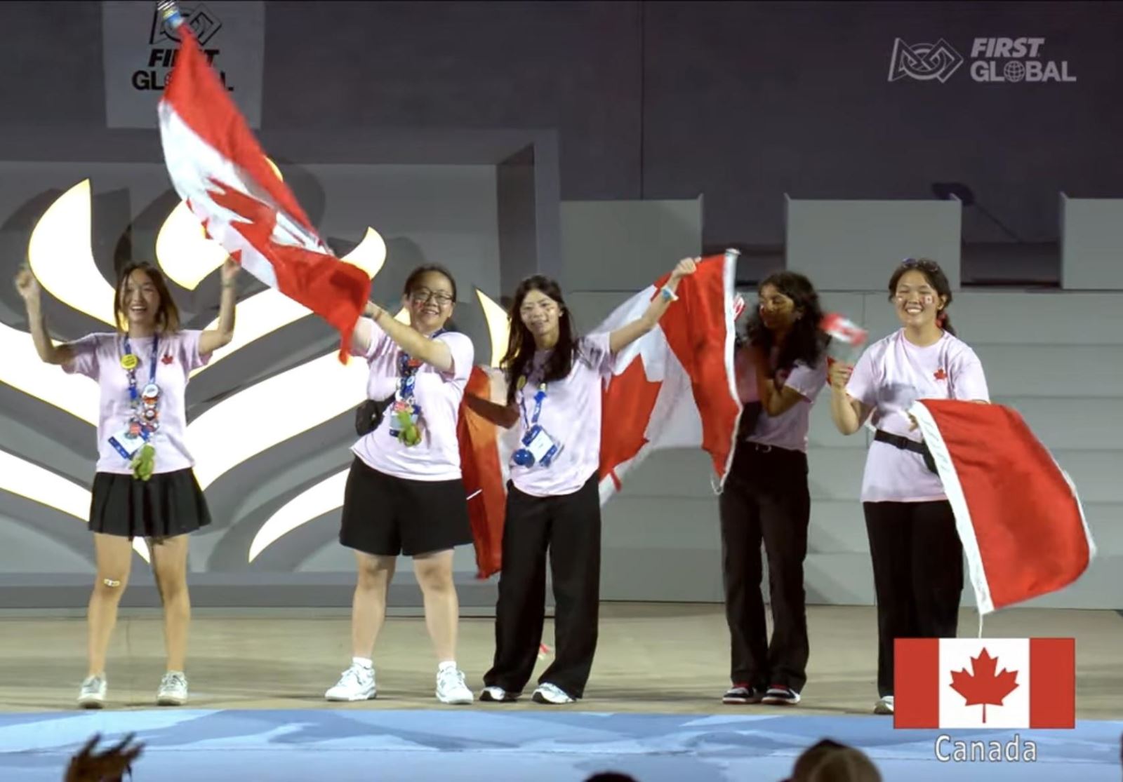 01 – Five student members from Team Canada stand on a stage waving Canadian flags during the opening ceremony at the FIRST Global Challenge in Athens, Greece.