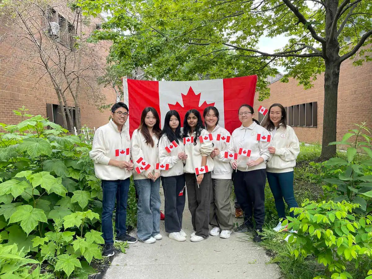 03 – Team Canada’s six student members and one staff advisor stand outside their school Dr. Norman Bethune CI in front of a Canadian flag and each hold two mini Canadian flags.