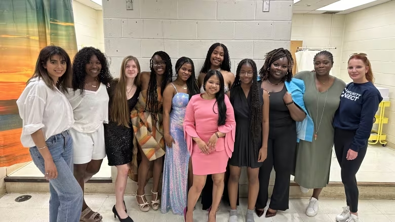 Six female students stand in a school hallway in formal dresses and surrounded by five women who are both stylists and staff who helped prepare the students for an evening at the awards event.