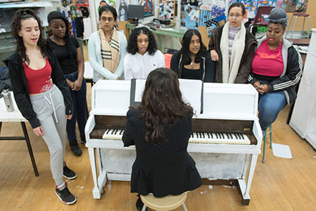 A teacher sits at a piano while seven students stand around it singing