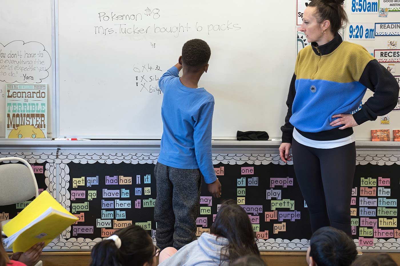 Student solves math problem on a whiteboard while educator and other students watch