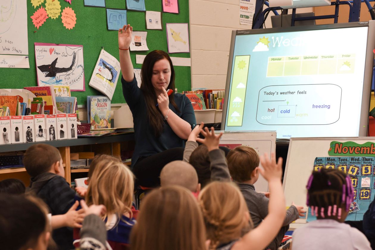 Teacher with hand raised teaching pronunciation with students seated on carpet