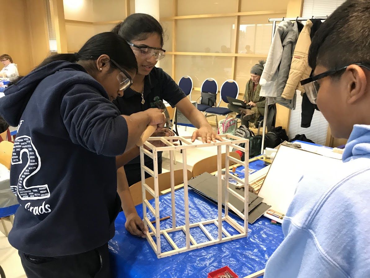 Three students wearing goggles construct wooden frame with hammer