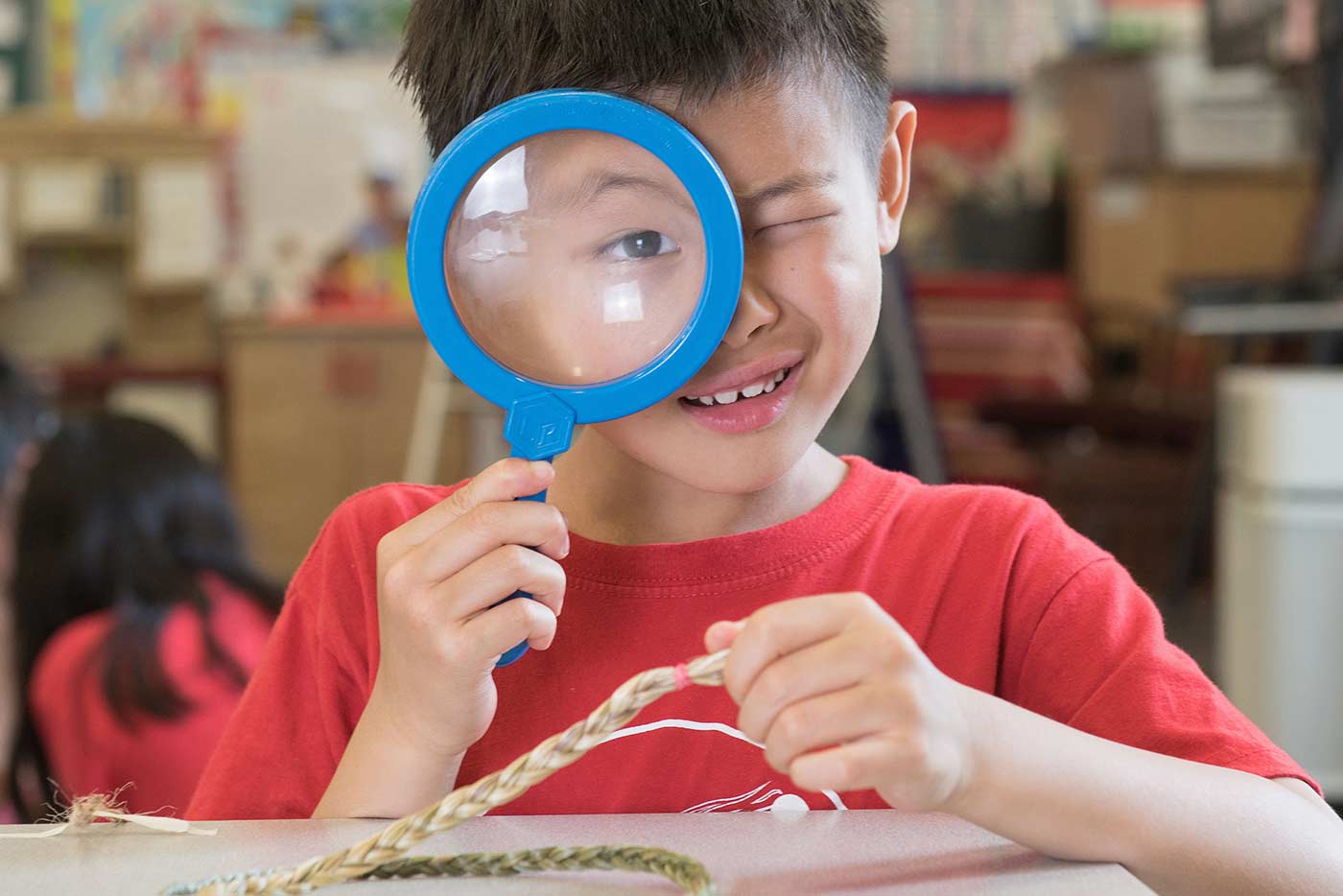 A young student wearing a red shirt examines a braided object through a magnifying glass